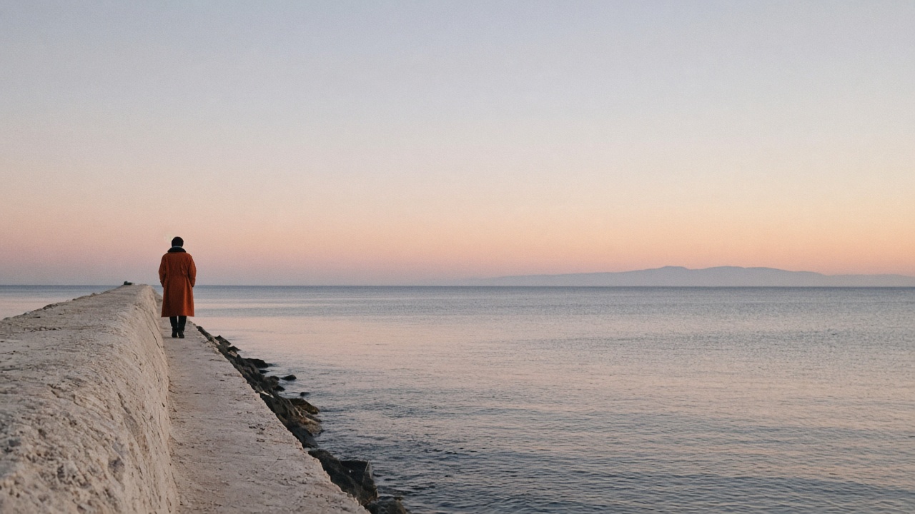 A person in a rust coat walking a sea wall, kept small on the left, with open sky and water filling most of the frame